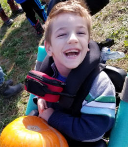 A boy is sitting in a specialized stroller with a neck harness and a pumpkin on his lap.