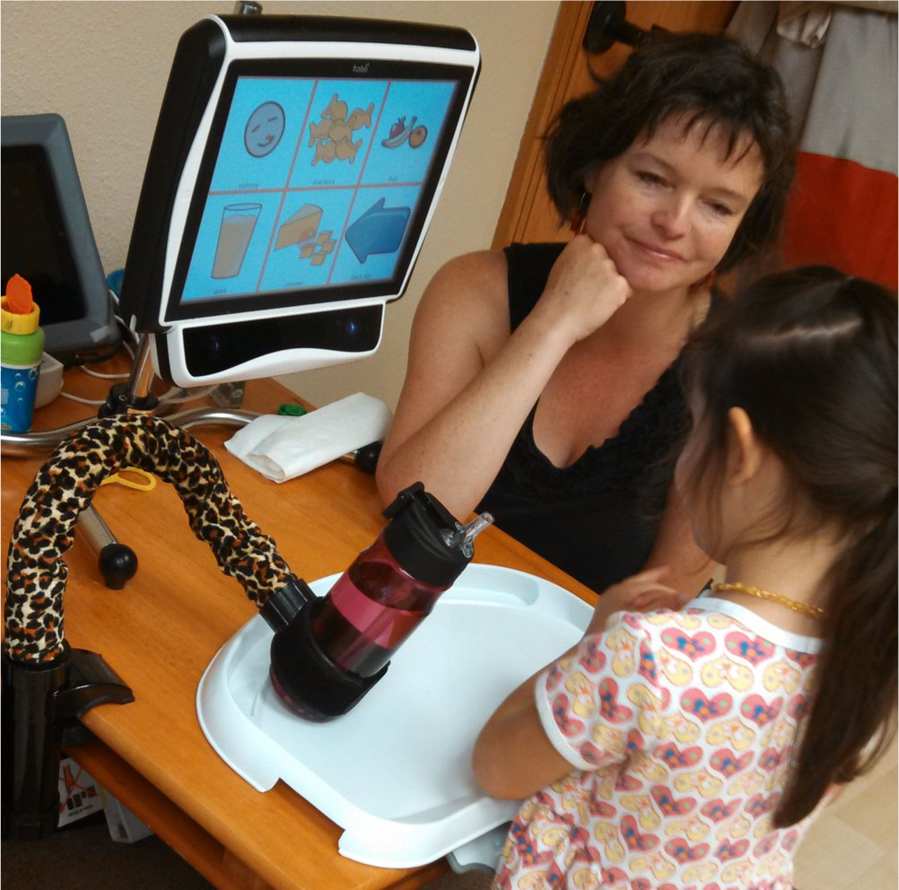 Little girl using her eye gaze speech generating device, with a woman watching her.