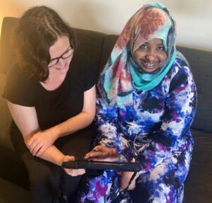 Two women sit on couch. One shows the other a tablet to use as a communication device.