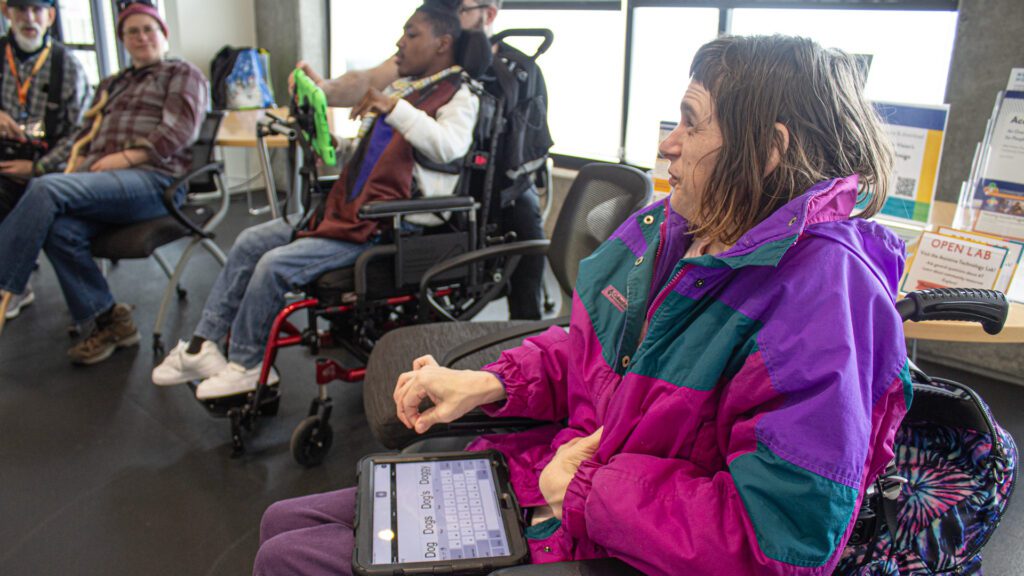 A woman smiles and she has a communication device on her lap. There are two other AAC user in the background.