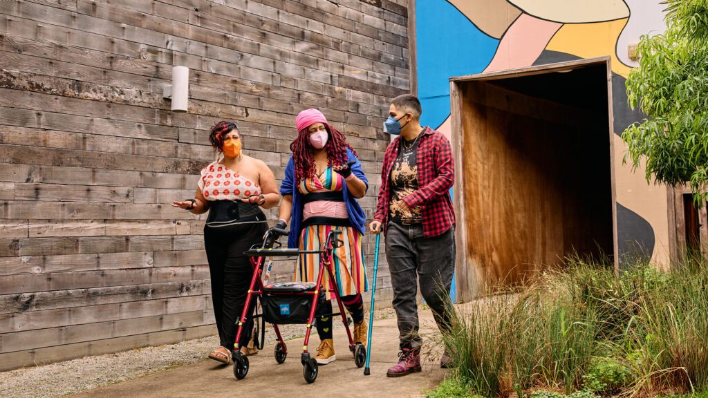 Three masked disabled people of color chat while strolling through an outdoor courtyard. On the left, an Indigenous woman wears glasses, wrist support braces, and waist trainers paired with a off-shoulder top and wide leg pants. In the middle, a Black non-binary woman walks with a rollator and wears waist trainers with ice pack inserts over their rainbow striped dress. On the right, a Latinx non-binary person walks while using a teal functional grip cane.